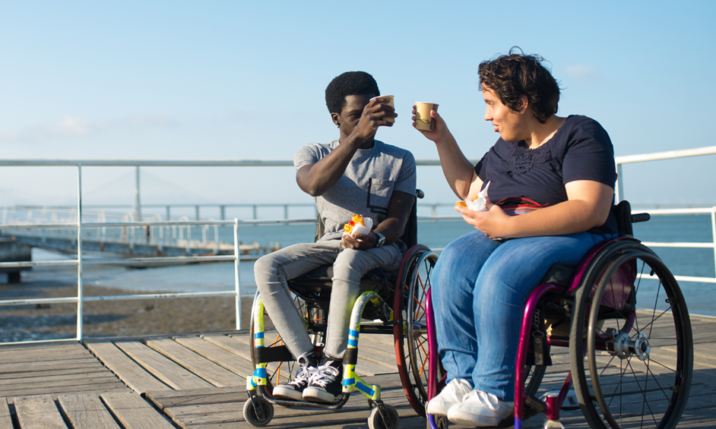 Two people cheersing their coffee while seated in their wheelchairs by the water