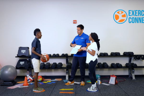 Three people standing in a room with exercise equipment around them. A young man is holding a basketball on the left. Coach Dave is showing him how many times to throw the ball on dry erase board. A young girl stands to the right in the photo waiting for the basketball to be thrown.