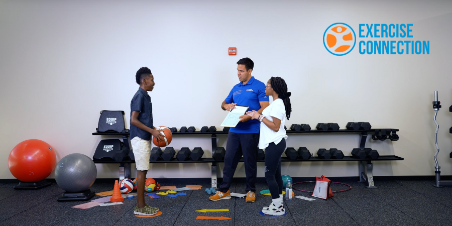 Three people standing in a room with exercise equipment around them. A young man is holding a basketball on the left. Coach Dave is showing him how many times to throw the ball on dry erase board. A young girl stands to the right in the photo waiting for the basketball to be thrown.