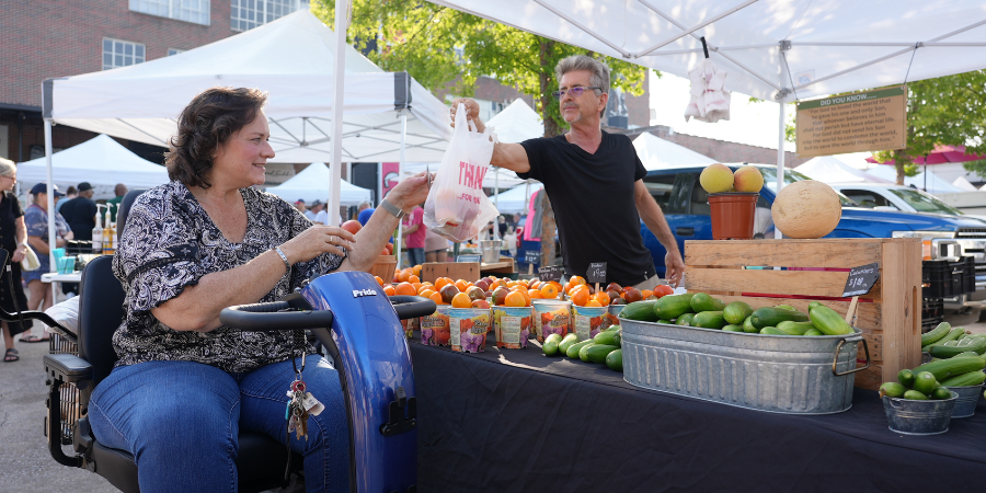 A woman using an electric cart to shop for fresh produce at a farmers market. A man is handing her a bag of produce.